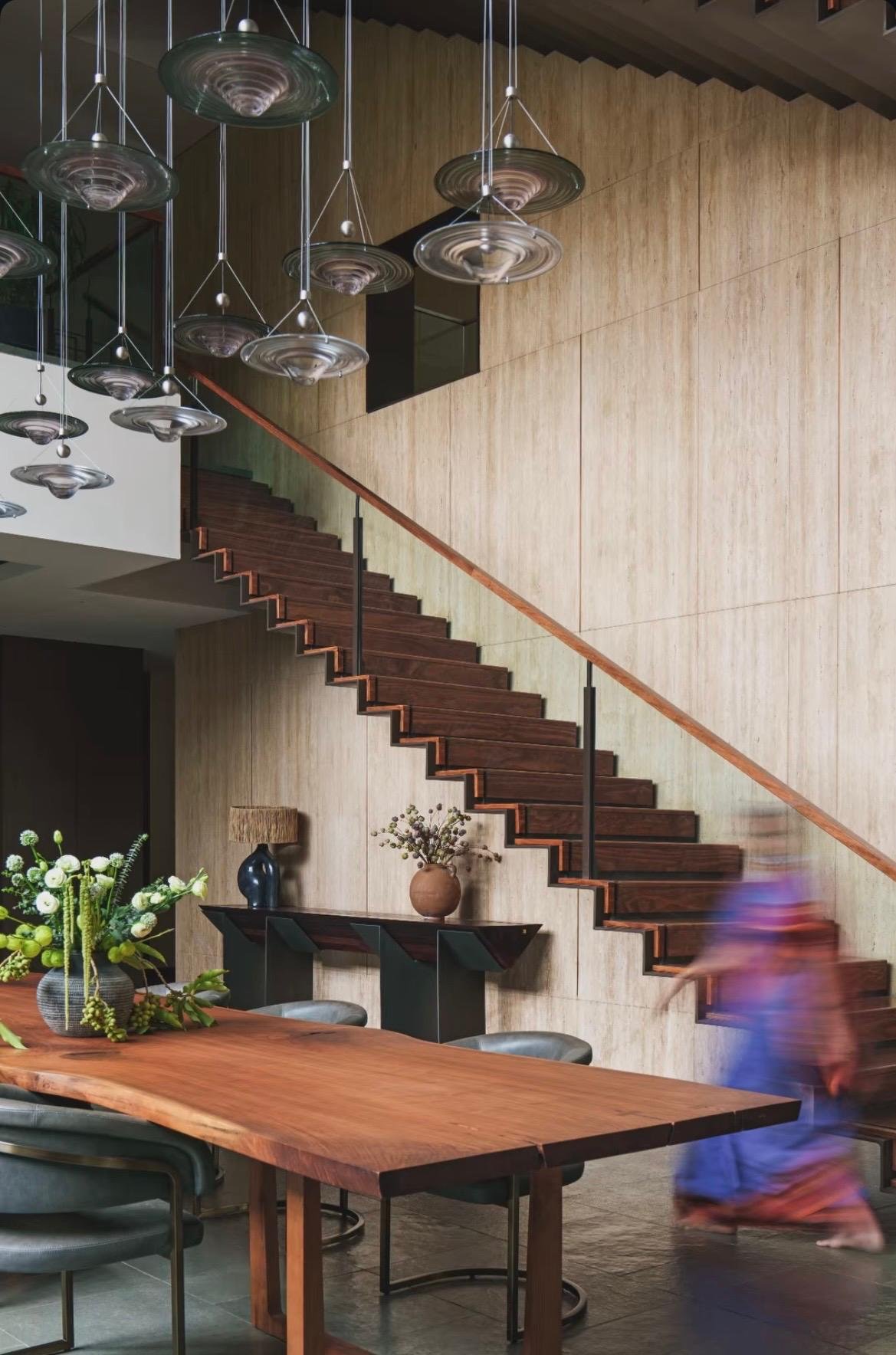 Dining space beneath glass pendants, with a timber staircase against a travertine wall
