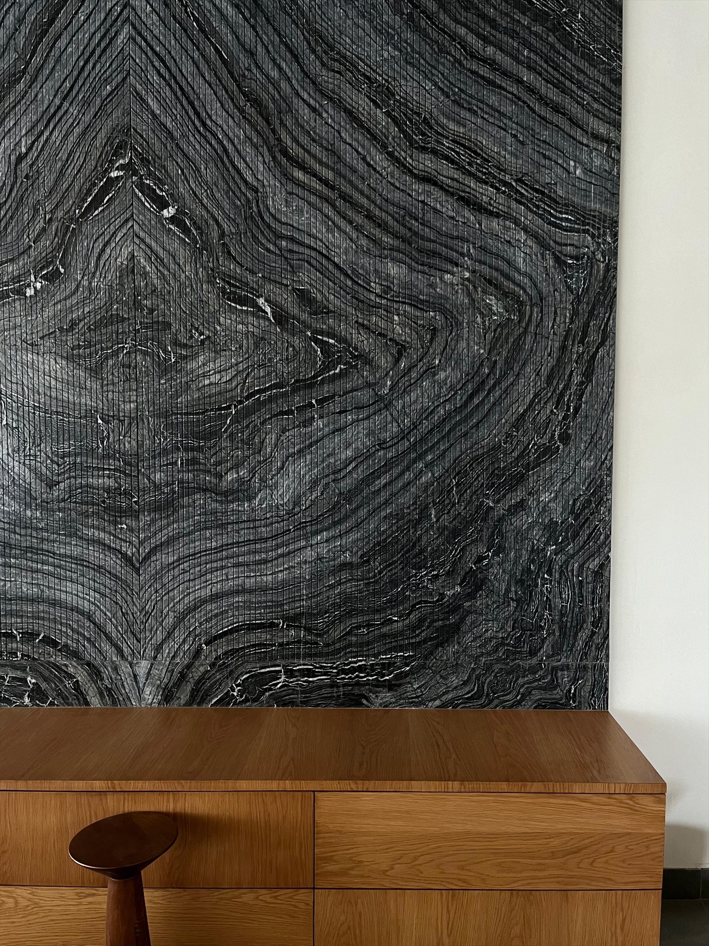 Book-matched fluted black marble wall above a low oak sideboard and a turned walnut stool