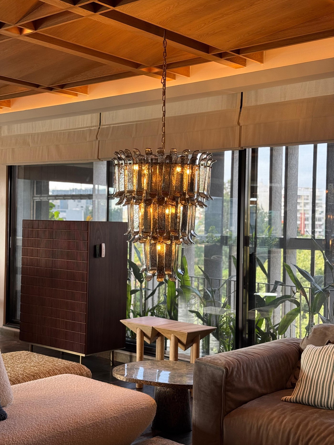 Smoked glass tiered chandelier beneath the coffered oak ceiling, with a fluted walnut cabinet and planted balcony beyond