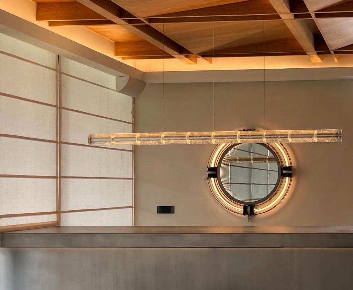 Linear crackle-glass pendant hung beneath the oak ceiling, before a backlit round mirror and linen blinds