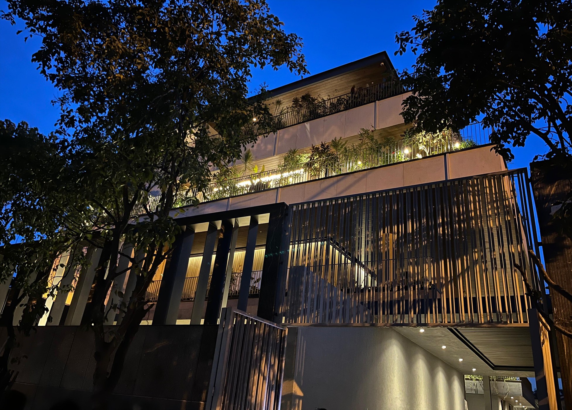 The Stepped Haven at dusk, with lit terraces and the vertical metal screen seen through the trees