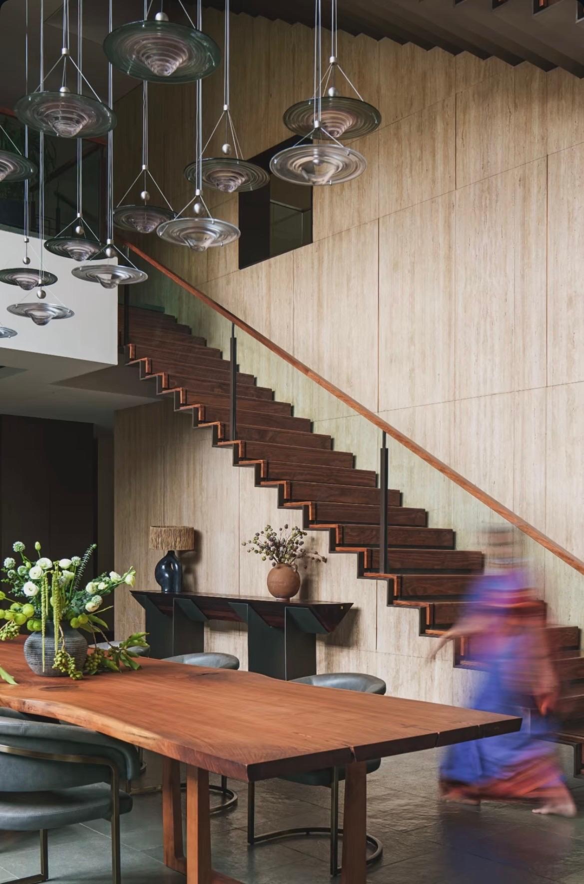 Double-height dining space at The Stepped Haven, with the timber cantilevered staircase against the stone wall and glass pendant lights above